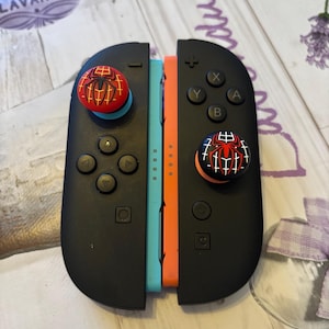 Op de afbeelding: Zwarte Nintendo Switch Joy-Con controllers met rood-zwarte Spider-Man duimgrepen. De controllers zijn gescheiden, met turquoise en oranje rails zichtbaar. De controllers hebben zwarte knoppen en richtingspads.