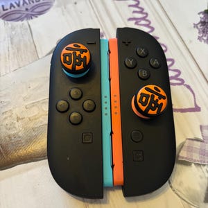 May include: Black Nintendo Switch Joy-Con controllers with orange and blue accents. The controllers have orange thumb grips with Japanese characters. The controllers are on a lavender and white patterned surface.