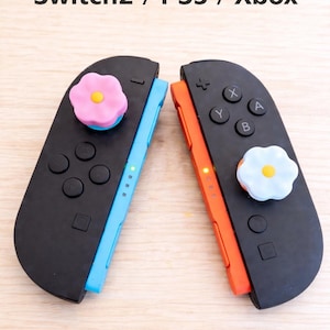 Puede incluir: Dos mandos de juego negros con laterales azules y naranjas, cada uno con un botón en forma de flor. El texto "Switch2 / PS5 / Xbox" está encima de los mandos. Los mandos tienen botones y luces indicadoras.