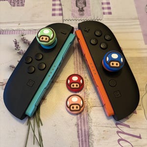 May include: Two black Nintendo Switch Joy-Cons with mushroom-shaped thumb grips. One Joy-Con has a teal side, the other is orange. Three additional mushroom thumb grips are also visible: red, pink, and green.