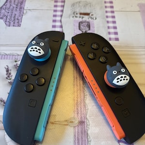 May include: Two black Nintendo Switch Joy-Cons with Totoro-themed thumb grips. One Joy-Con has a turquoise side, the other has an orange side. The buttons are black, and the thumb grips are grey and white.