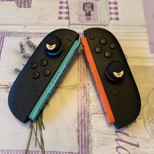 May include: Two black Nintendo Switch Joy-Con controllers with Batman-themed thumbstick caps. One controller has a teal side, the other orange. The controllers are on a patterned surface.