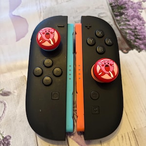 May include: Black Nintendo Switch Joy-Con controllers with blue and orange sides. Each controller has a red and pink Kirby-themed joystick cap. The controllers are on a patterned surface.