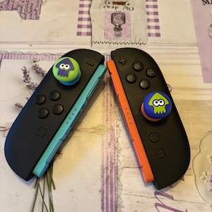 May include: Two black Nintendo Switch Joy-Con controllers with colorful accents. One has a teal side and a green and purple squid-shaped thumb grip. The other has an orange side and a blue and green squid grip.