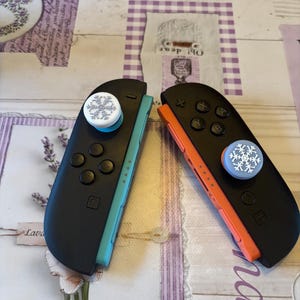 May include: Two black game controllers with snowflake thumb grips. One controller has a teal side and the other has an orange side. The controllers are resting on a lavender and white patterned surface.