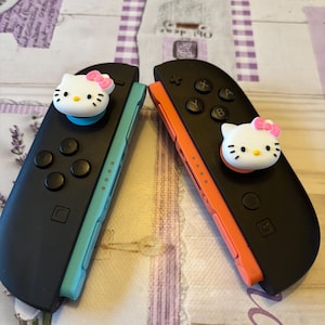 May include: Two black Nintendo Switch Joy-Con controllers, one with a teal side and the other with an orange side. Each controller has a white Hello Kitty face with a pink bow on the joystick.