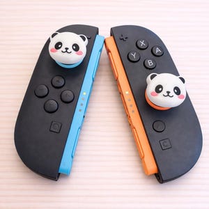 May include: Two black game controllers with blue and orange accents, each topped with a white panda face. The controllers have buttons and directional pads. Text at the top reads "Switch2 / PS5 / Xbox".