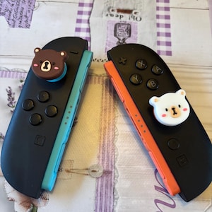 May include: Two black Nintendo Switch Joy-Con controllers with bear-shaped thumb grips. One controller has a teal side, the other has an orange side. The bear grips are brown and white, adding a cute touch to the gaming accessories.
