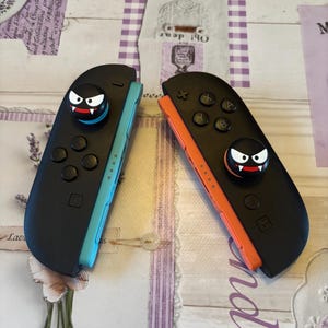 May include: Two black game controllers with a cartoon monster face on the joystick. One controller has a blue side, and the other has an orange side. The controllers are on a patterned surface.