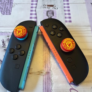 May include: Two black Nintendo Switch Joy-Con controllers with red and yellow Manchester United logo thumb grips. One controller has a teal side, the other has an orange side. Buttons are black.