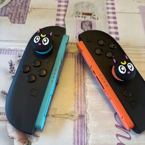 May include: Two black game controllers, each with a cartoon cat face on the joystick. One controller has a turquoise side, the other an orange side. They rest on a patterned surface.