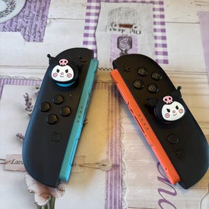 May include: Two black game controllers with colorful accents. One controller has a teal side, the other has an orange side. Each controller has a black character charm with a skull and face design. The controllers are on a patterned surface.