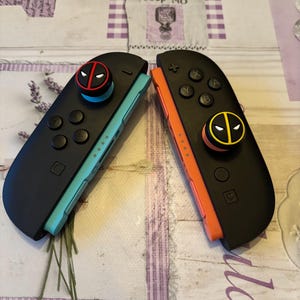 May include: Two black Nintendo Switch Joy-Con controllers, one with a teal side and the other with an orange side. Each has a Deadpool-themed button. The controllers are on a patterned surface.