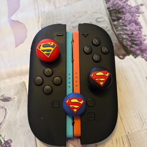 May include: Black Nintendo Switch Joy-Con controllers with red, blue, and black Superman logo thumb grips. The controllers have blue and orange wrist straps. The background is a light purple and white floral pattern.