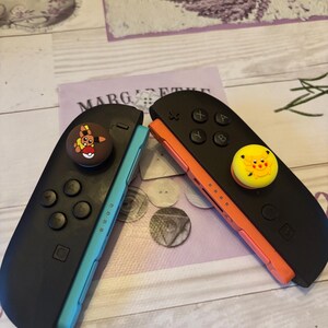May include: Two black Nintendo Switch Joy-Con controllers, one with a teal side and the other with an orange side. Each controller has a decorative button on top. One button features a cartoon character, and the other has a yellow face.