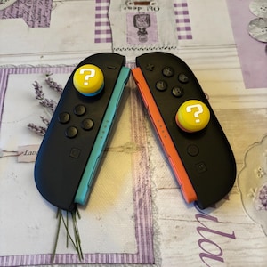 May include: Two black Nintendo Switch Joy-Con controllers. One has a teal side and the other has an orange side. Each controller has a yellow joystick with a white question mark. The controllers are on a lavender floral patterned surface.