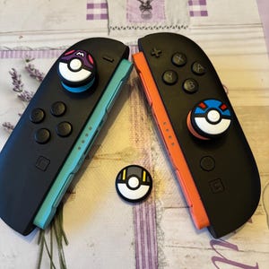 May include: Two black Nintendo Switch Joy-Cons with colorful accents, featuring custom thumb grips with a Pokemon theme. One Joy-Con has a teal side and the other has an orange side. A third thumb grip is also visible.