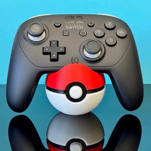 May include: A black Nintendo Switch Pro Controller with white detailing, placed on a red, white, and black Poké Ball. The controller features buttons marked X, Y, A, and B. The background is a solid blue.