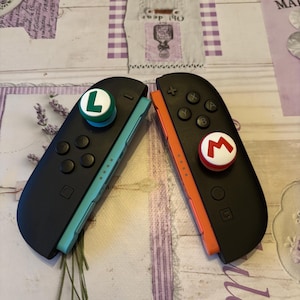 May include: Two black Nintendo Switch Joy-Con controllers, one with a green "L" button and the other with a red "M" button. The controllers have black buttons and are set against a lavender floral background.