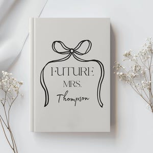 May include: A pale grey book with the text "FUTURE MRS. Thompson" in black script. A black bow design is above the text. The book is surrounded by white flowers and a white cloth.