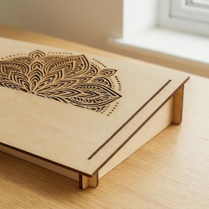 Könnte beinhalten: Eine hellfarbene Holzkiste mit einem dekorativen, geätzten Blumendesign auf dem Deckel. Die Kiste ist abgewinkelt, mit einer erhöhten Rückseite und einer niedrigeren Vorderseite. Das Design ist ein detailliertes, symmetrisches Muster. Die Kiste wird wahrscheinlich zur Aufbewahrung oder Präsentation verwendet.