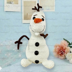 May include: A white crocheted snowman plush toy featuring a bright orange carrot nose, two black eyes, a wide stitched smile, and three black buttons down the front. The figure has brown twig-like arms and a brown branch-like hair piece on top.