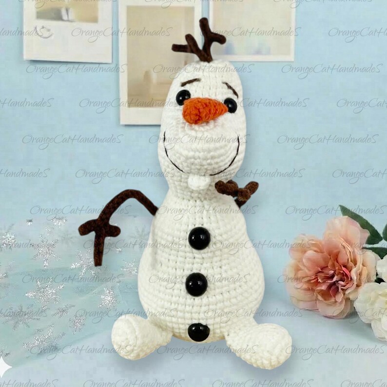 May include: A white crocheted snowman plush toy featuring a bright orange carrot nose, two black eyes, a wide stitched smile, and three black buttons down the front. The figure has brown twig-like arms and a brown branch-like hair piece on top.