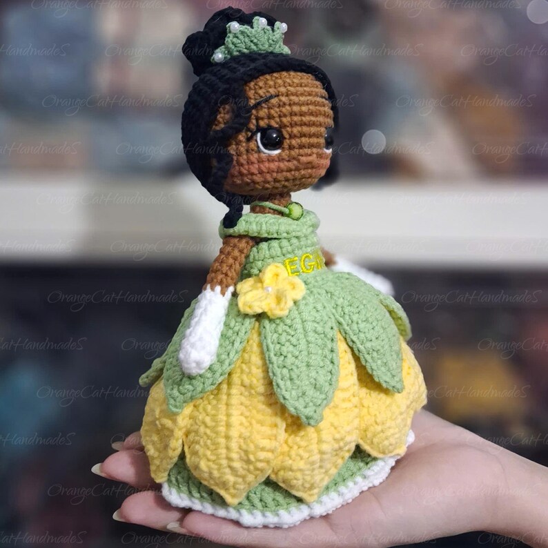 May include: A handmade crochet doll of a princess in a green and yellow dress. The doll has black hair, a green crown, and white gloves. A yellow flower is on the dress. The doll is held in a hand.