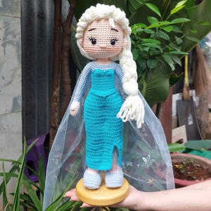 May include: A handmade crochet doll of a character with a white braided hairstyle, wearing a blue dress with a slit. The doll has a light blue cape and is mounted on a wooden base. The doll is approximately 12 inches tall.