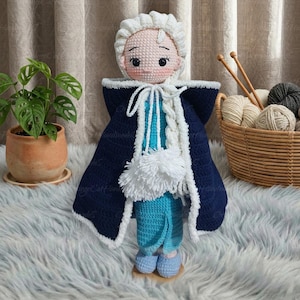 May include: A handmade crochet doll in a blue and white outfit, featuring a white hood and cape. The doll has a light pink face with black eyes and a white braid. The doll is standing on a wooden base.