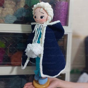 May include: A handmade crochet doll depicting a character in a navy blue cape with white trim. The doll has a white wig, a light blue dress, and blue shoes. A white pom-pom hangs from the cape. The doll is displayed on a wooden base.