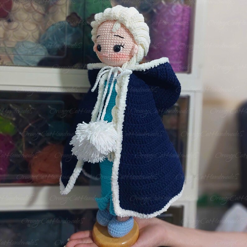 May include: A handmade crochet doll depicting a character in a navy blue cape with white trim. The doll has a white wig, a light blue dress, and blue shoes. A white pom-pom hangs from the cape. The doll is displayed on a wooden base.