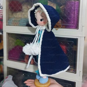 May include: A handmade crochet doll wearing a navy blue hooded cape with white trim and pom-poms. The doll has a light skin tone, white hair, and is wearing blue shoes. The doll is standing on a wooden base.