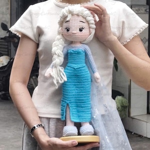 May include: A handmade crochet doll resembling a princess, featuring a light blue dress with a slit, long white braid, and light blue shoes. The doll is held on a wooden base.
