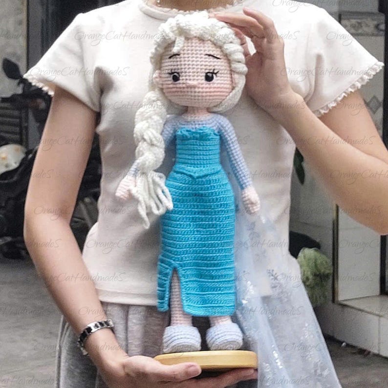 May include: A handmade crochet doll resembling a princess, featuring a light blue dress with a slit, long white braid, and light blue shoes. The doll is held on a wooden base.