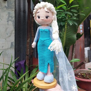 May include: A handmade crochet doll of a character with a light skin tone, wearing a turquoise dress with long sleeves and a white cape. The doll has a white braided hairstyle and is standing on a wooden base. The doll is approximately 10 inches tall.