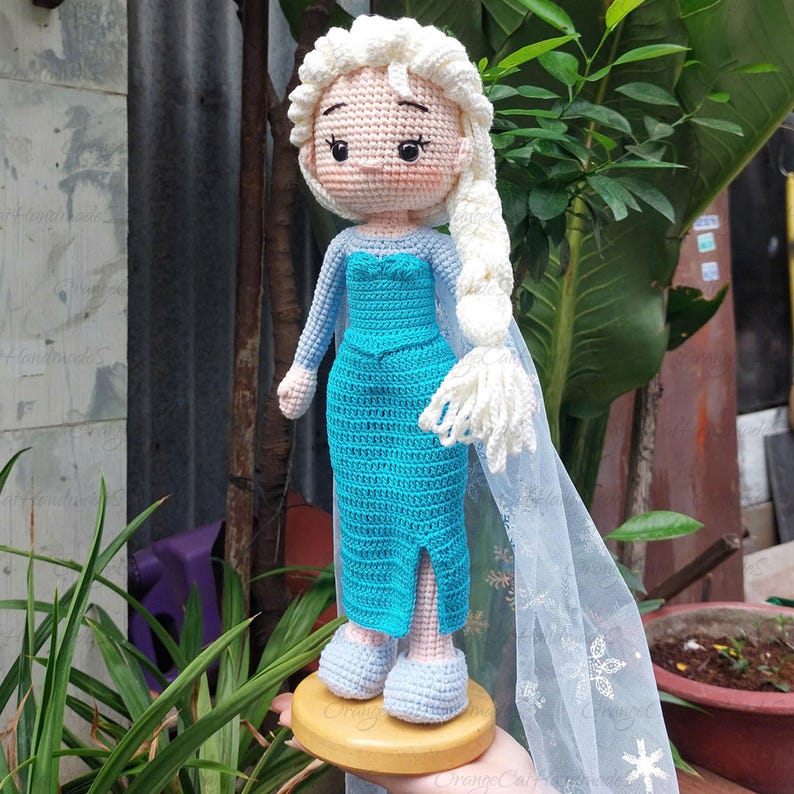 May include: A handmade crochet doll of a character with a light skin tone, wearing a turquoise dress with long sleeves and a white cape. The doll has a white braided hairstyle and is standing on a wooden base. The doll is approximately 10 inches tall.
