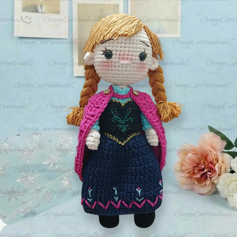 May include: A handcrafted crochet doll featuring light brown yarn hair styled in two braids. The doll wears a dark blue dress with embroidered floral details and a bright pink cape. It has a round head with black eyes and rosy cheeks.