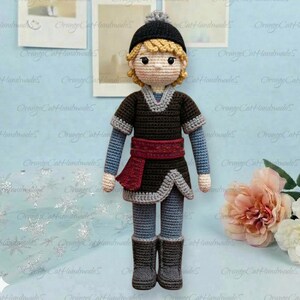 May include: A handmade crochet doll featuring a person with blonde hair wearing a black beanie, a dark brown tunic with grey trim, a red sash, grey pants, and dark grey boots. The doll has a light skin tone and is crafted with yarn.