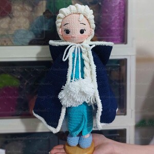 May include: A handmade crochet doll featuring a light skin tone, rosy cheeks, and a serene expression. The doll wears a blue and white dress with a dark blue cape and a white braided hairstyle. The doll is approximately 8 inches tall.