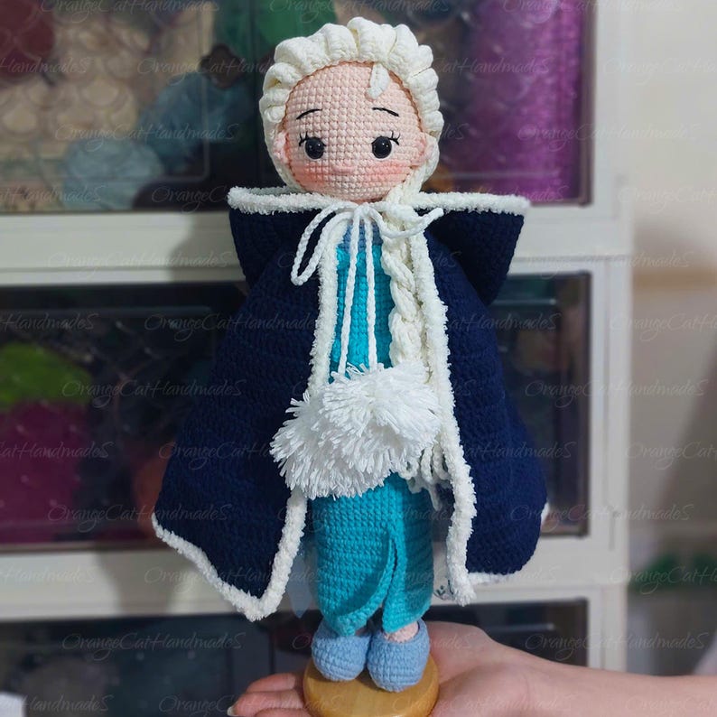 May include: A handmade crochet doll featuring a light skin tone, rosy cheeks, and a serene expression. The doll wears a blue and white dress with a dark blue cape and a white braided hairstyle. The doll is approximately 8 inches tall.