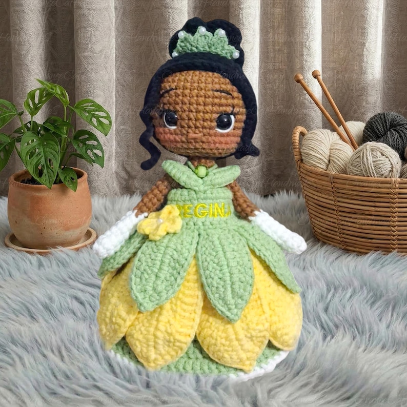 May include: A handmade crochet doll of a princess, featuring a brown complexion, black hair, and a green and yellow dress. The doll has a flower detail and the word "EGIN" on the dress. The doll is sitting on a fluffy, gray surface.