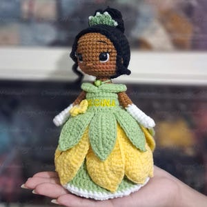 May include: A handmade crochet doll of a princess, featuring a black bun, a green crown, and a green and yellow petal-shaped dress. The doll has brown skin, large eyes, and white gloves. The name "RECINA" is embroidered on the dress.