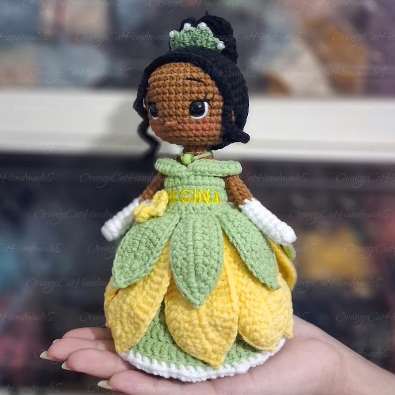 May include: A handmade crochet doll of a princess, featuring a black bun, a green crown, and a green and yellow petal-shaped dress. The doll has brown skin, large eyes, and white gloves. The name "RECINA" is embroidered on the dress.