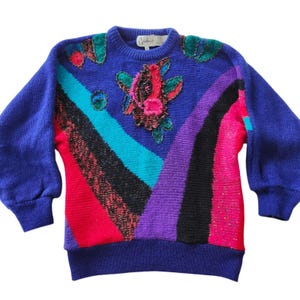 Può includere: Un maglione blu vintage con girocollo e maniche lunghe. Il maglione presenta un design colorato con strisce rosse, nere, turchesi e viola. È impreziosito da applicazioni floreali e a forma di foglia.