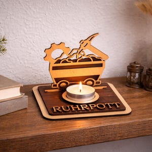 Könnte beinhalten: Kerzenhalter aus Holz mit einem Grubenwagen-Design. Der Kerzenhalter zeigt ein Zahnrad, eine Spitzhacke und das Wort "RUHRPOTT". Eine brennende Teelichtkerze befindet sich in der Mitte. Der Artikel ist aus hellem und dunklem Braunholz.