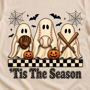 May include: A cream-coloured t-shirt featuring a graphic of three ghosts wearing baseball-themed sheets, holding a glove, a baseball, and bats. Below them are two jack-o'-lanterns, a checkered border, and the text 'Tis The Season in black font.