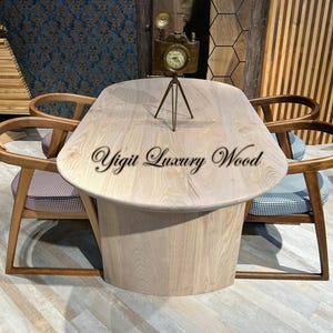 May include: A light-colored wooden table with a unique, rounded shape, paired with wooden chairs featuring patterned cushions. A vintage-style clock in the shape of a camera sits on the table. The text "Yigit Luxury Wood" is on the table.
