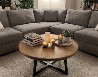 Könnte beinhalten: Ein runder Couchtisch aus Holz mit naturfarbener Oberfläche und einem schwarzen, X-förmigen Metallgestell. Auf der Tischplatte befinden sich ein Bücherstapel, mehrere zylindrische Kerzen in Gläsern, eine kleine Grünpflanze und eine Schale.