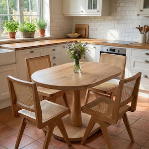 May include: A light wood oval dining table with four matching chairs. The chairs have woven cane backs and seats. The kitchen has white cabinets, a white tile backsplash, and a window with plants.